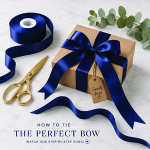 Kraft gift box wrapped with navy satin ribbon tied into a perfect gift bow beside a ribbon roll and gold scissors.